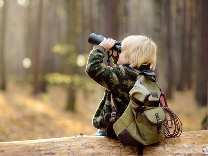 Ein Kind sitzt auf einem Baumstamm im Wald und schaut durch ein Fernglas. 