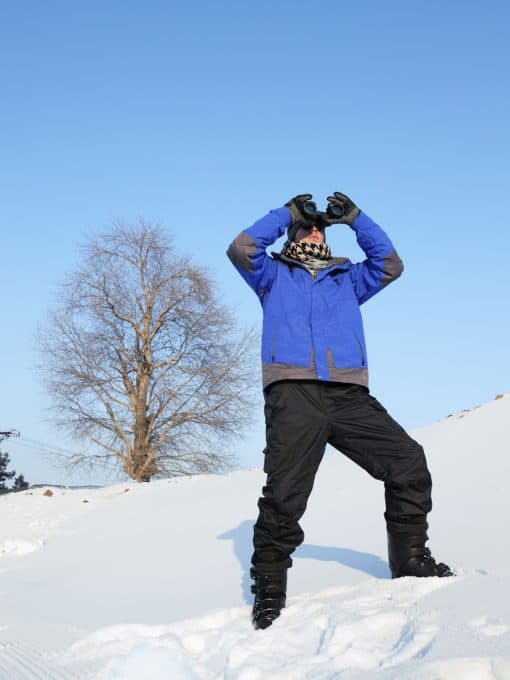 Ein Mann steht mit Skischuhen im Schnee und schaut mit dem Fernglas in die Ferne.